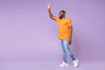 Wall Mural - Full length side view of joyful young african american man wearing basic casual orange t-shirt waving and greeting with hand as notices someone isolated on pastel violet background, studio portrait.