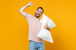 © ViDi Studio - Joyful young bearded man in pajamas home wear hold pillow clenching fist doing winner gesture while resting at home isolated on yellow background studio portrait. Relax good mood lifestyle concept.