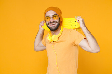 Cheerful smiling young bearded man 20s wearing basic casual t-shirt headphones eyeglasses hat standing hold skateboard looking camera isolated on bright yellow colour background, studio portrait.