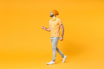 Wall Mural - Full length side view of cheerful smiling funny young man 20s wearing basic casual t-shirt headphones hat walking going looking aside isolated on bright yellow colour background, studio portrait.