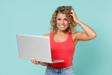 Preoccupied young blonde woman 20s in pink basic casual tank top standing working on laptop pc computer put hand on head looking camera isolated on blue turquoise colour background studio portrait.