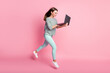 Full size profile photo of cheerful pretty brunette girl run jump writing laptop wear shirt pants sneakers isolated on pink color background