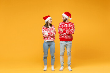 Wall Mural - Full length of smiling young Santa couple friends man woman in sweater Christmas hat hold hands crossed looking at each other isolated on yellow background. Happy New Year celebration holiday concept.