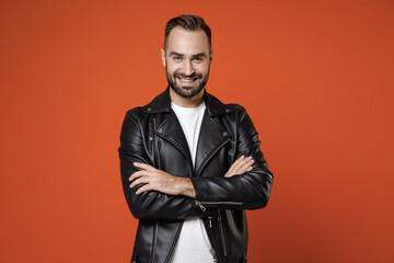 Wall Mural - Smiling confident handsome young bearded man 20s wearing basic white t-shirt black leather jacket standing holding hands crossed looking camera isolated on orange colour background studio portrait.