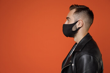 Wall Mural - Side view of young bearded man 20s wearing white t-shirt black leather jacket face mask safe from coronavirus virus covid-19 during pandemic quarantine isolated on orange background studio portrait.