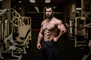  strong young adult bearded male with perfect sport muscle physique in dark night indoors gym among white exercise equipment