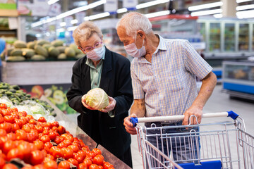 mature couple in mask and gloves with covid protection picks tomatoes in vegetable section of supermarket