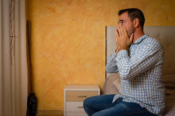 Attractive bearded man depressed and upset in the bedroom. Dramatic 30-40 year old lifestyle portrait, handsome man sitting on bed, worried and desperate