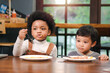 © Stella - Cute African American boy with curly and adorable Asian kid eating meal at the table indoor, happy children having food in a restaurant, enjoy their meal together