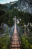 Fototapeta Sypialnia - Wooden long footbridge above the deep gorge with a river beach at the bottom, between the rocks. Wild mountains in Swiss Alps