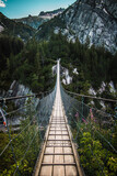 Fototapeta Mosty linowy / wiszący - Wooden long footbridge above the deep gorge with a river beach at the bottom, between the rocks. Wild mountains in Swiss Alps