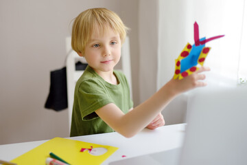 Boy making colored paper crafts at home during coronavirus lockdown. Child showing result teacher by laptop. Creativities lessons, distance learning, homeschooling, online kindergarten for kids.