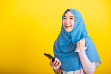 Wall Mural - Asian Muslim Arab, Portrait of happy beautiful young woman Islam religious wear veil hijab funny smile she raise hand glad celebrating winning with smart mobile phone isolated on yellow background