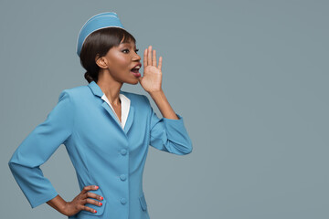 Wall Mural - Cheerful young african stewardess speaks into empty space. Blue background.