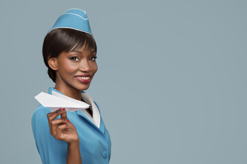 Wall Mural - Smiling young african stewardess holding a paper airplane in her hand. Blue background.