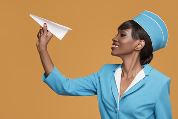 Wall Mural - Smiling young african stewardess holding a paper airplane in her hand. Orange background.