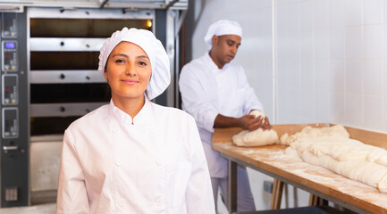 Wall Mural - Portrait of smiling young hispanic female professional baker in white uniform standing in bakery during working day