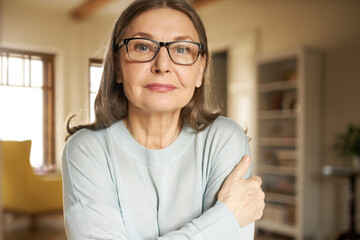 Horizontal portrait of senior gray haired European female in eyeglasses posing indoors, looking at camera with serious facial expression. Mature people, aging, retirement and lifestyle concept