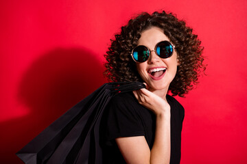 Photo of sweet cheerful wavy lady dressed casual black outfit eyewear holding back shopping bags looking empty space isolated red color background