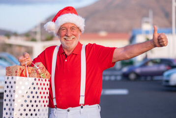 A happy senior bearded man shopping for Christmas in a Santa hat and suspenders, holding a shopping bag full of gifts and making the ok sign with his hand