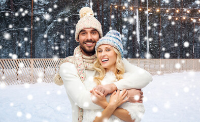 Poster - winter holidays, christmas and people concept - smiling man and woman in hats and scarf hugging over ice skating rink background