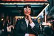 Millennial woman in trendy glasses with neon reflection looking up during evening sightseeing in New York, attractive brunette hipster girl dressed in stylish clothing enjoying travel time