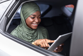 Wall Mural - Cheerful Black Muslim Woman In Hijab Using Digital Tablet While Ridind Car