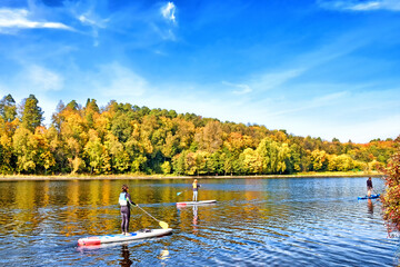 Wall Mural - golden autumn landscape with people enjoying water sport activity on river against scenic blue sky with clouds background. Colorful forest and river at city park. Perfect day of fall season