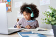 © insta_photos - Cute smiling african school kid girl wearing headphones virtual distance learning online listening remote education digital class doing homework studying at home classroom sitting at desk with laptop.