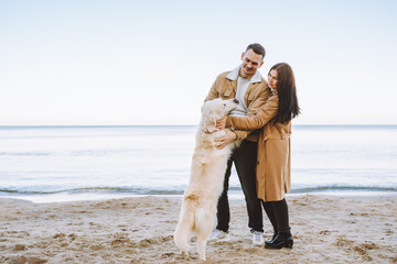 Family of young woman and man walking with their pet golden retriever by seaside at autumn sunny day. Back view