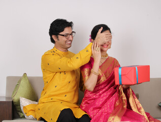 Indian husband surprising wife with a gift on Diwali / anniversary / birthday while sitting on sofa.