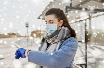 Wall Mural - health, safety and pandemic concept - young woman wearing protective medical mask looking at her wristwatch at bus stop in city over snow