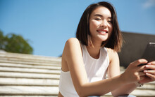 Image Of Beautiful Stylish Woman Sitting On Street Stairs On Summer Day And Holding Mobile Phone. Asian Girl Texting Message On Smartphone And Smiling At Camera