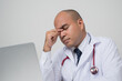 © Chanakon - A portrait of Asian doctor put on stethoscope tired rubbing nose and eyes feeling fatigue and headache sitting at the desk isolated on white background.