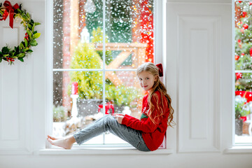 Girl with red bow sitting on the big window with snowing background.