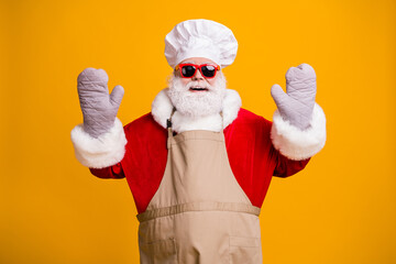 Poster - Photo of excited cheerful santa grandpa grey beard chef headwear bake cookery raise hands pot holders gloves wear red costume coat gloves sun specs cap apron isolated yellow color background