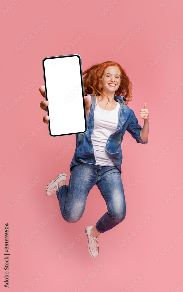 Happy redhead girl jumping with smartphone and showing thumb up - obrazy, fototapety, plakaty 