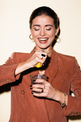 Wall Mural - happy woman in brown blazer squeezing lemon in glass with alcohol cocktail on white