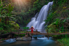 A Young Asian Tourist Wearing A Red Dress Stands On A Bamboo Bridge Watching Pha Dok Siew Waterfall At Doi Inthanon National Park, Chiang Mai Province, Thailand.