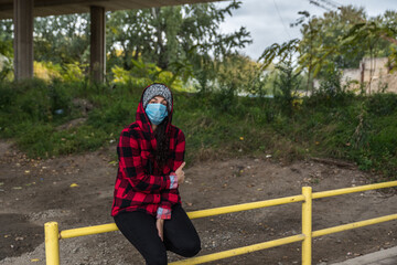 Wall Mural - Young depressed homeless girl or woman with hood and protective medicine mask sitting outdoor scared from coronavirus virus or COVID-19 feeling abandoned and anxious selective focus