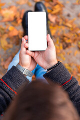 Man holding smartphone with white screen outdoors with yellow autumn leaves on the ground. Empty display for copy space