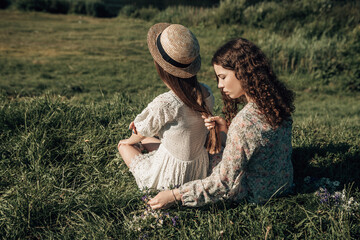 Back view of two young ladies sitting on the grass in the sunny morning summer country, looking at the river or lake. Girls dressed in bright dress braids hair, nature background copy space.
