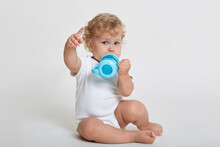 Portrait Of Cute Toddler Drinking Water From Blue Bottle Cup, Indicating Away With His Index Finger, Little Charming Male With Blond Curly Hair Looking Directly At Camera Isolated On White Background.