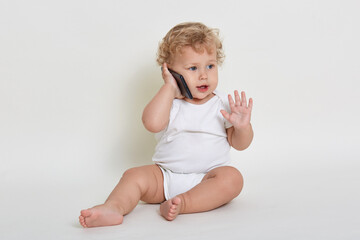 Cute and joyful baby boy playing talking with somebody via smartphone while sitting indoors on floor, looking away and raising palm up, wearing white body suit, play with mobile phone.