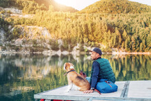 Man Dog Owner And His Friend Beagle Dog Are Sitting On The Wooden Pier On The Mountain Lake And Enjoying The Landscape During Their Walking In The Autumn Season Time. Human And Pet Concept Image.
