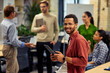 © Friends Stock - Young cheerful mixed race man holding digital tablet and smiling at camera while working with colleagues in the modern office, selective focus