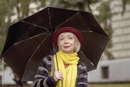 Street portrait of an elderly woman 60-65 years old in a hat under an umbrella on the background of the city landscape. Concept: retirement, fashionista of advanced years, excellent health.