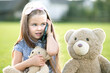 © bilanol - Pretty child girl sitting in summer park on green grass with her teddy bear toy talking on mobile phone outdoors in summer.