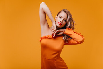 Wall Mural - Amazing slim girl in orange dress dancing in studio. Happy curly female model posing on yellow background