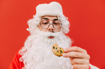 Close portrait of a man in a suit of Santa Claus on a red background, looking hungry at the chocolate chip cookies in his hand. Christmas and New Year.